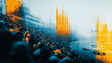 Crowded grandstand overlooks racetrack with two racing cars, overlaid with blue and orange pixelated digital effects.